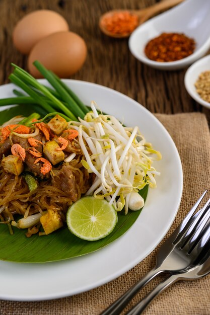 Pad Thai en un plato blanco con limón, huevos y condimentos en una mesa de madera.