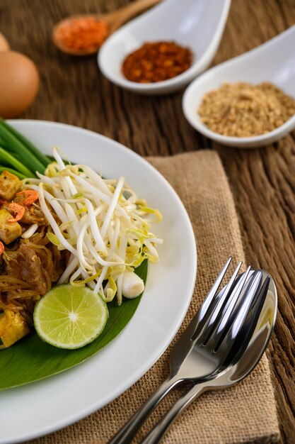 Pad Thai en un plato blanco con limón, huevos y condimentos en una mesa de madera.