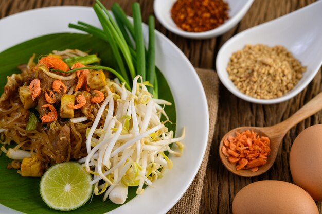 Pad Thai en un plato blanco con limón, huevos y condimentos en una mesa de madera.