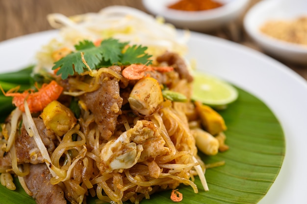 Pad Thai en un plato blanco con limón, huevos y condimentos en una mesa de madera.