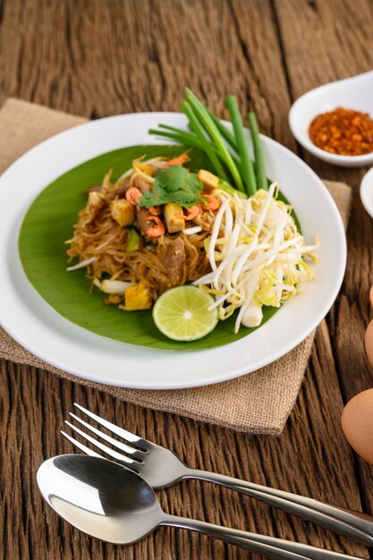 Pad Thai en un plato blanco con limón, huevos y condimentos en una mesa de madera.