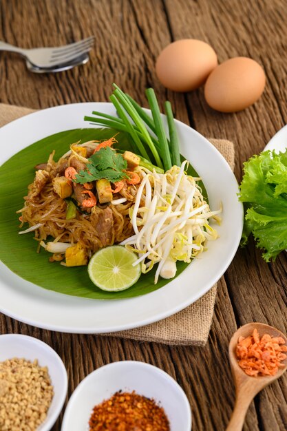 Pad Thai en un plato blanco con limón, huevos y condimentos en una mesa de madera.