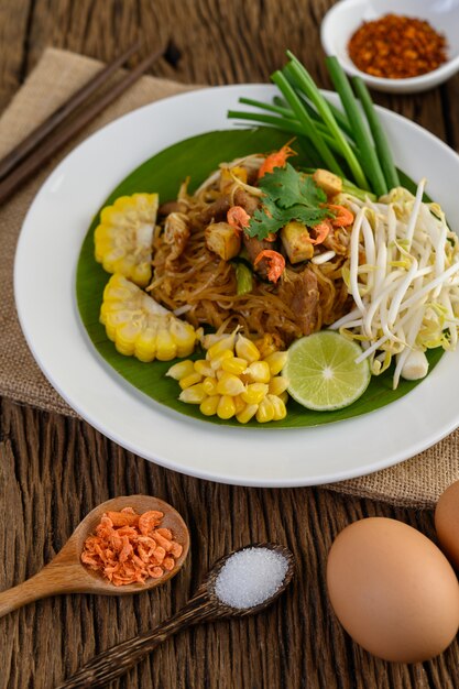 Pad Thai en un plato blanco con limón, huevos y condimentos en una mesa de madera.