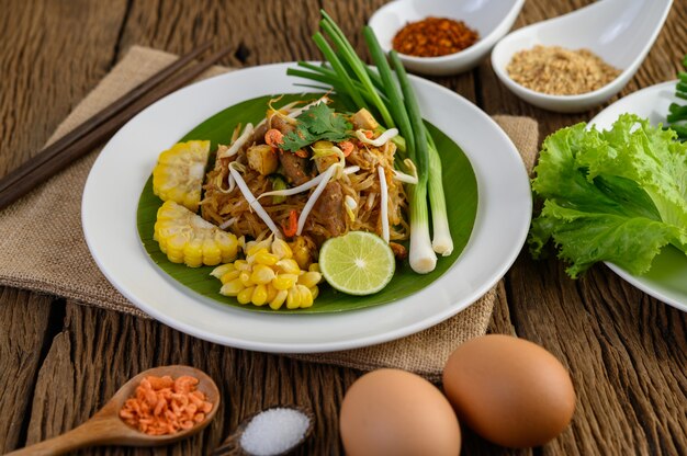 Pad Thai en un plato blanco con limón, huevos y condimentos en una mesa de madera.