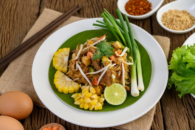 Pad Thai en un plato blanco con limón, huevos y condimentos en una mesa de madera.
