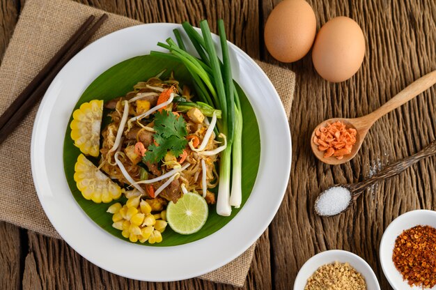 Pad Thai en un plato blanco con limón, huevos y condimentos en una mesa de madera.