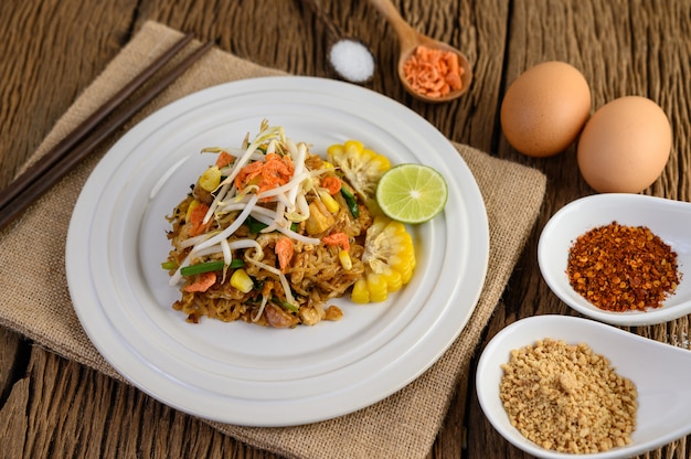 Pad Thai en un plato blanco con limón, huevos y condimentos en una mesa de madera.