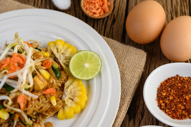 Pad Thai en un plato blanco con limón, huevos y condimentos en una mesa de madera.