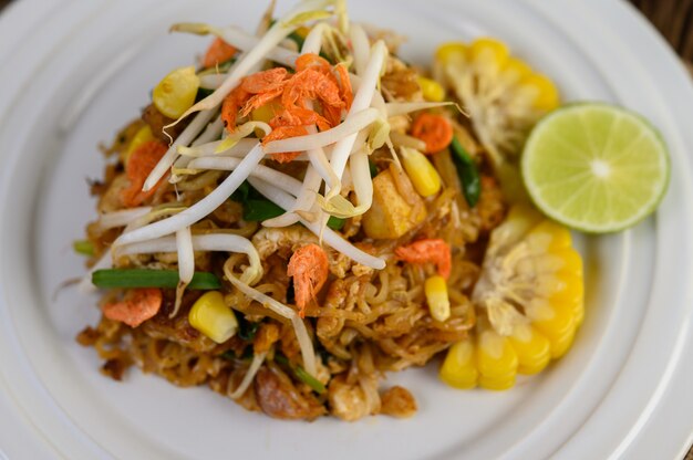 Pad Thai en un plato blanco con limón, huevos y condimentos en una mesa de madera.