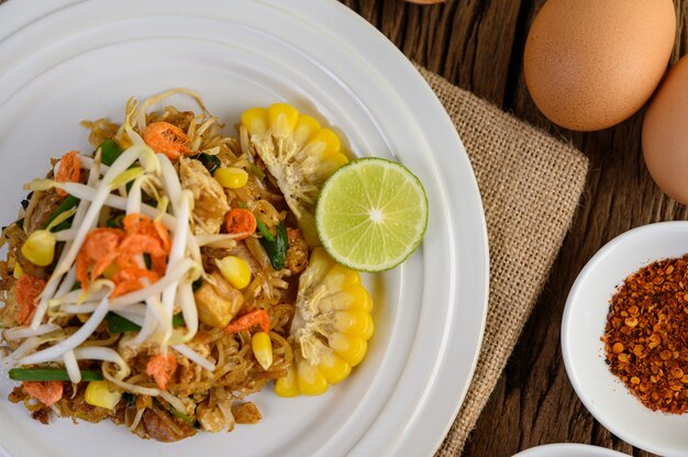 Pad Thai en un plato blanco con limón, huevos y condimentos en una mesa de madera.