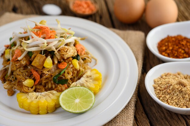 Pad Thai en un plato blanco con limón, huevos y condimentos en una mesa de madera.