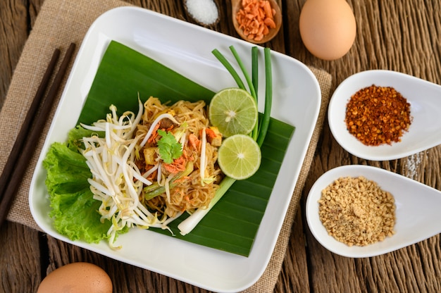 Pad Thai en un plato blanco con limón, huevos y condimentos en una mesa de madera.