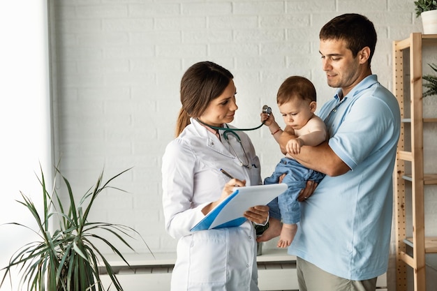 Padre adulto medio sosteniendo a su hijo pequeño y comunicándose con una doctora que está tomando notas en el portapapeles