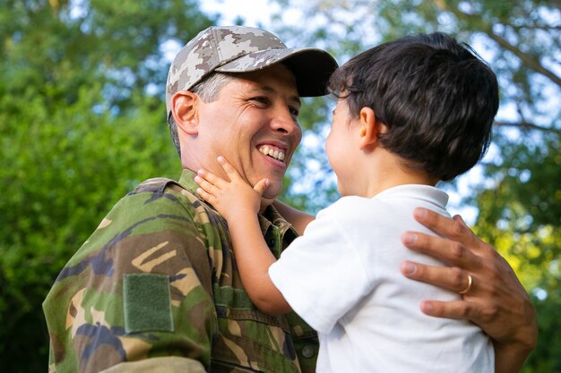 Padre alegre sosteniendo a su pequeño hijo en brazos, abrazando al niño al aire libre después de regresar de un viaje misionero militar Ángulo bajo. Reunión familiar o concepto de regreso a casa