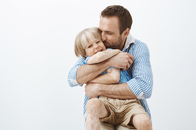 El padre ama a su pequeño hijo como nadie. Lindo y guapo papá cariñoso abrazando y besando al niño en la mejilla, sintiéndose feliz de pasar tiempo con el niño, de pie sobre una pared gris