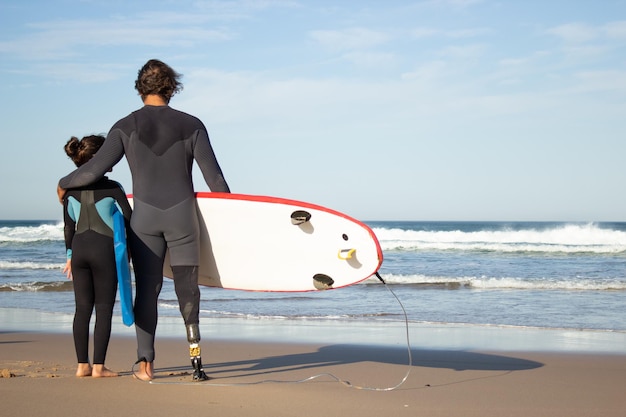 Padre amoroso con pierna mecánica con hija en la playa. Vista posterior de un hombre adulto medio y una niña de cabello oscuro que llevan tablas de surf, mirando el agua. Familia, ocio, concepto de estilo de vida activo.