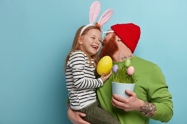 El padre cariñoso besa a la hija y se sostiene de las manos, sostiene una olla de huevos decorados, se prepara para la Pascua Feliz niña de jengibre lleva orejas de conejo, lleva un gran huevo amarillo. Fiestas religiosas, concepto de celebración