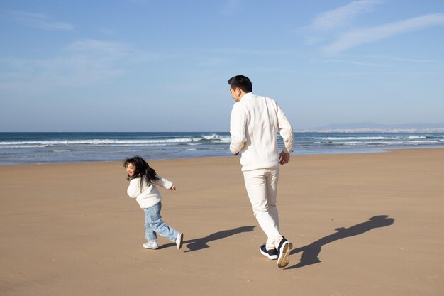 Padre e hija activos jugando en la playa. Familia japonesa corriendo tras la otra en un día soleado. Papá ocio, tiempo en familia, concepto de crianza