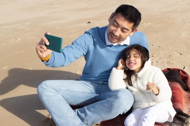 Padre e hija japoneses complacidos tomando selfie en la playa. Niño de cabello oscuro y hombre sosteniendo teléfono, haciendo muecas. Familia, concepto de tecnología