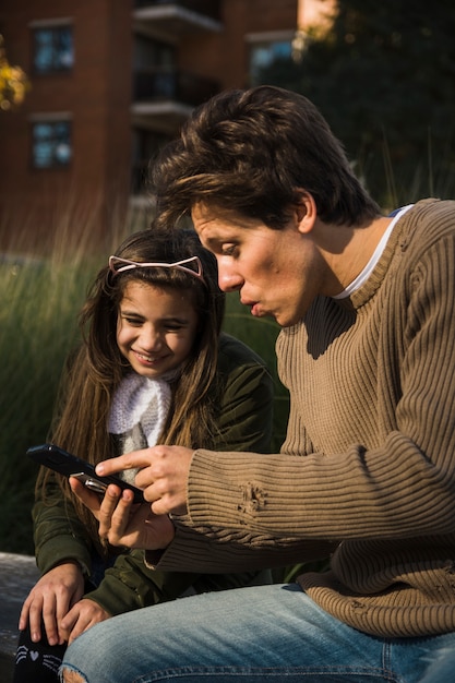 Foto gratuita padre e hija mirando la pantalla del teléfono móvil mientras está sentado en el parque