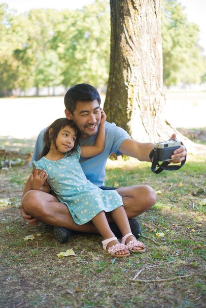 Padre e hijo asiáticos jóvenes tomando sus fotos en cámara. Hombre sonriente sentado en el suelo con una hija pequeña de rodillas y tomando fotos mirando a la cámara. Concepto de ocio y paternidad