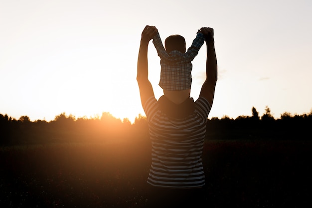 Padre e hijo caminando en el campo a la hora de la puesta del sol, muchacho sentado en los hombros de los hombres