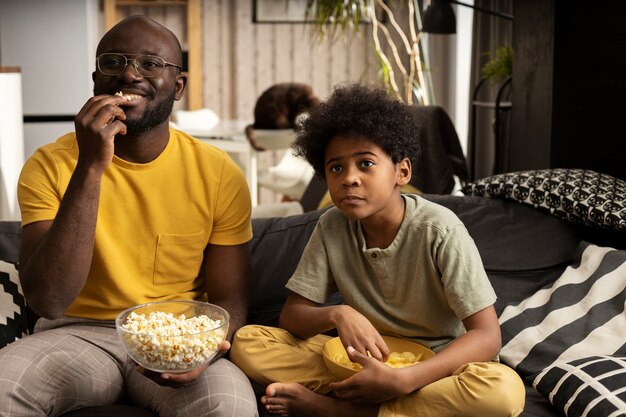 Padre e hijo con palomitas de maíz y papas fritas juntos en el sofá