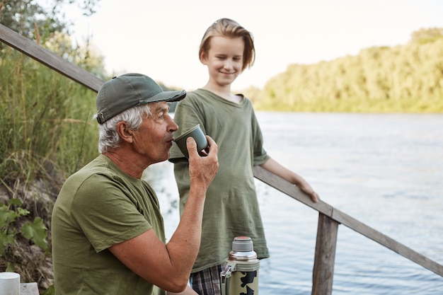 Foto gratuita padre e hijo pescando en la orilla del río o lago, hombre mayor bebiendo té del termo, familia posando en las escaleras de madera que conducen al agua, descansar en la hermosa naturaleza.