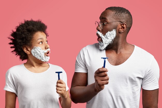 Padre e hijo sorprendidos con crema de afeitar en la cara, sujetan las navajas, sorprendidos porque no tienen tiempo para la higiene matutina