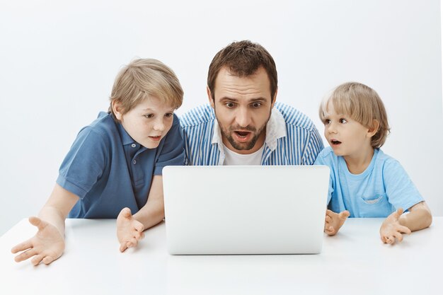 Padre e hijos viendo video tutorial, queriendo sorprender a la madre. Retrato de papá y niños confundidos despistados sentados en la mesa y mirando la pantalla del portátil