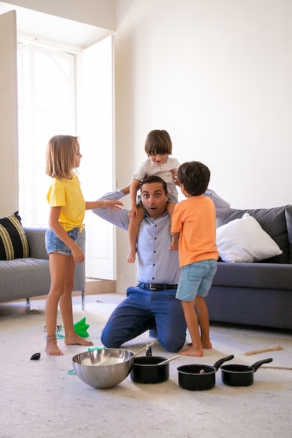 Padre emocionado divirtiéndose con los niños en la sala de estar. Papá feliz con hijo sobre los hombros. Adorable niña y niño de pie cerca de ellos. Sartenes y cuenco para caza. Concepto de infancia, fin de semana y hogar.