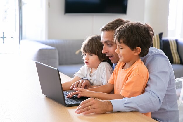 Padre emocionado mostrando algo en la computadora portátil a los hijos pequeños. Adorables niños caucásicos que aprenden computadora en casa con la ayuda de un amoroso papá de mediana edad. Concepto de paternidad, infancia y tecnología digital.