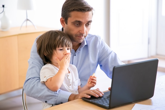 Padre enfocado usando laptop e hijo de rodillas. Papá de mediana edad caucásico sentado a la mesa con el niño pequeño lindo y trabajando en la computadora. Concepto de infancia, freelance y paternidad