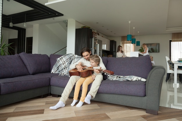 Padre enseñando a su hijo tocar guitarra, hija ayudando a su madre en la cocina