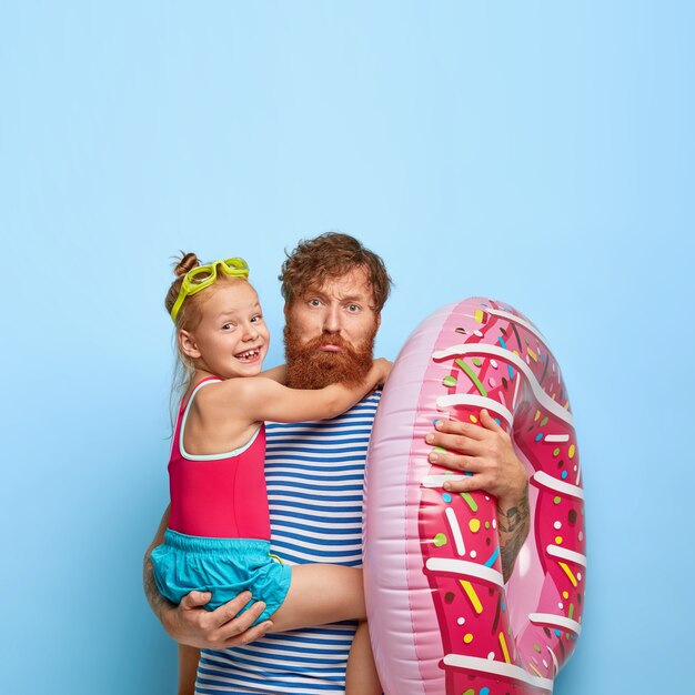 Padre de fatiga triste con barba pelirroja, lleva a su hija pequeña en las manos, traje de baño inflado, van a la playa juntos, vestidos de manera informal, entretener en el lugar de descanso