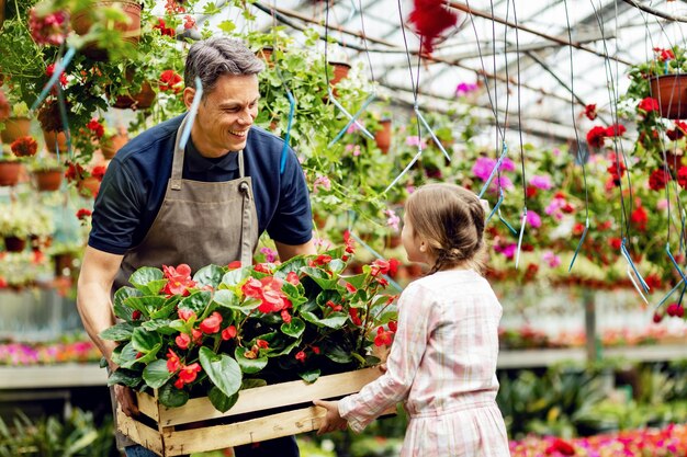 Padre feliz cargando una caja con flores mientras su pequeña hija lo ayuda en el centro de jardinería