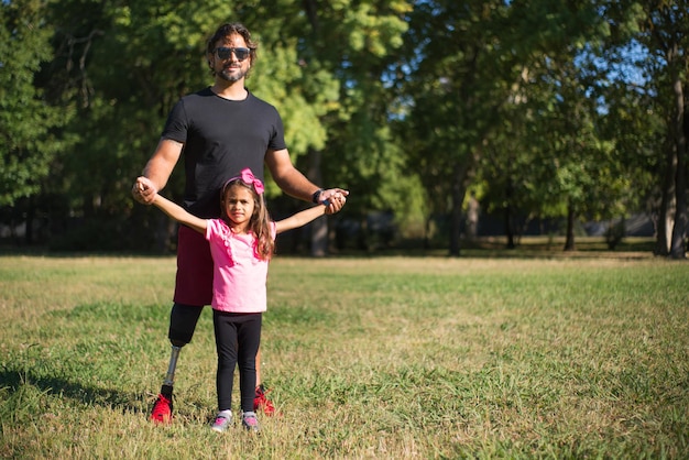 Padre feliz con discapacidad con hija. Hombre con pierna mecánica en gafas de sol y linda niña en el parque, tomados de la mano. Discapacidad, familia, concepto de amor