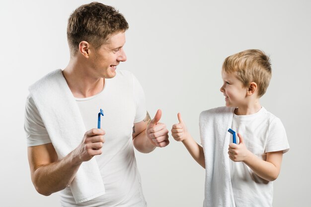 El padre y el hijo sonrientes que sostienen la maquinilla de afeitar en la mano que muestra el pulgar para arriba firman en el fondo blanco contra el contexto blanco