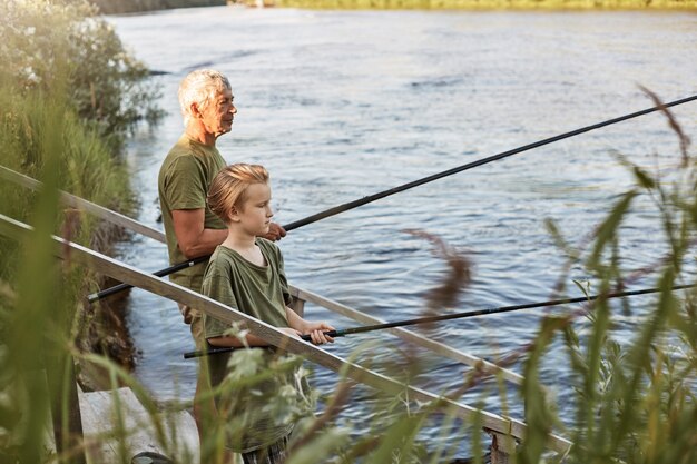 Padre maduro de pelo gris europeo con hijo pescando al aire libre en el lago o río, de pie cerca del agua con cañas de pescar en las manos, vestido informal, disfrutando de la afición y la naturaleza.