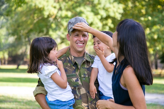 Padre militar alegre en uniforme que regresa a la familia, sosteniendo a dos niños en brazos. Mujer ajuste casquillo de los maridos. Reunión familiar o concepto de regreso a casa