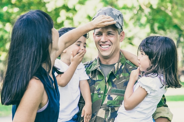 Padre militar alegre en uniforme regresando a la familia, sosteniendo a dos niños en brazos