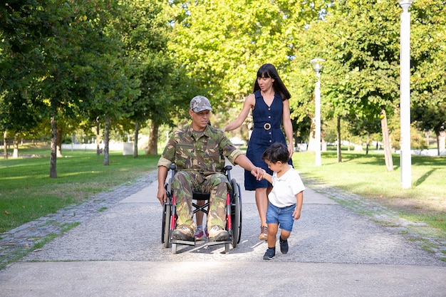 Padre militar serio caminando en silla de ruedas con la familia. Papá de mediana edad caucásico en uniforme de camuflaje sosteniendo la mano del hijo y hablando con una esposa bonita. Veterano de guerra y concepto de discapacidad