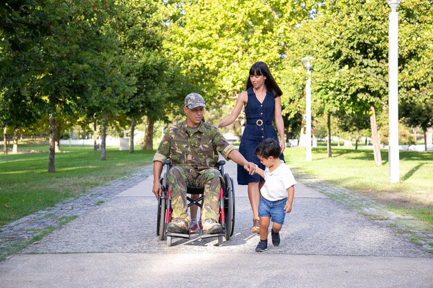 Padre militar serio caminando en silla de ruedas con la familia. Papá de mediana edad caucásico en uniforme de camuflaje sosteniendo la mano del hijo y hablando con una esposa bonita. Veterano de guerra y concepto de discapacidad