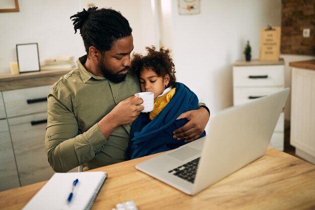 Padre negro cariñoso dando una taza de té a su pequeña hija enferma en casa