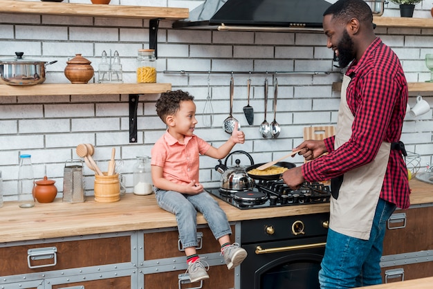 Padre negro cocinando comida mientras hijo mostrando pulgar arriba