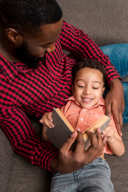 Padre negro y libro de lectura lindo del hijo en el sofá