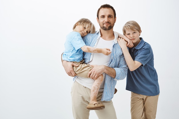 Padre soltero con dos niños lindos de pie. Papá sosteniendo a su hijo pequeño en las manos y sonriendo ampliamente mientras el hermano mayor se inclina sobre el hombro