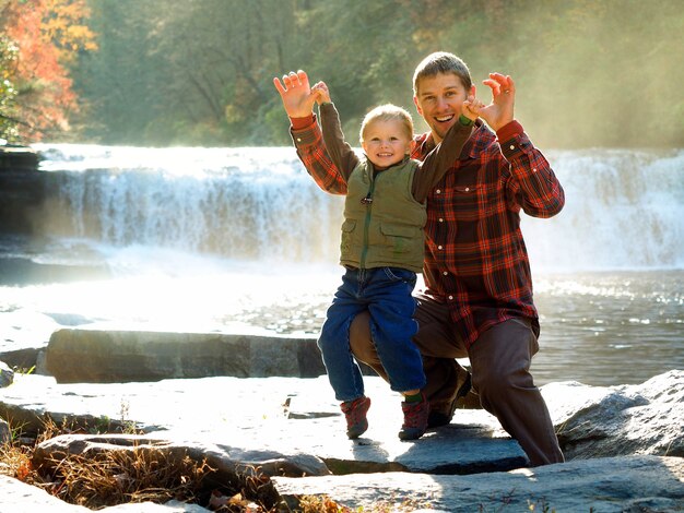 Padre sonriente con su hijo en un parque rodeado de vegetación y una cascada bajo la luz del sol