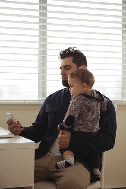 Padre sosteniendo a su bebé mientras usa el teléfono móvil en el escritorio