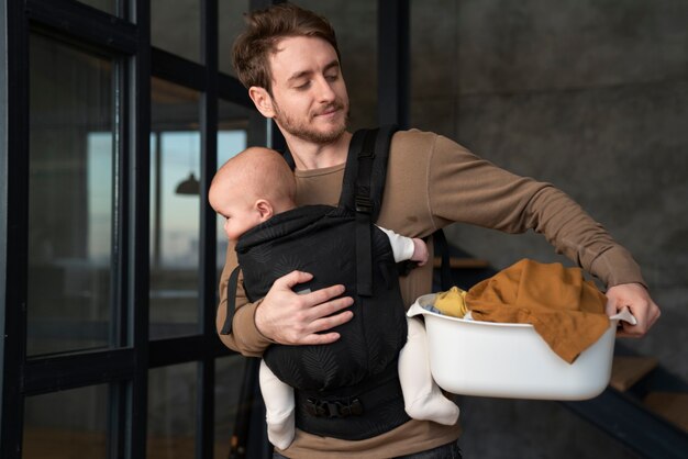 Padre de tiro medio sosteniendo al bebé y haciendo las tareas del hogar